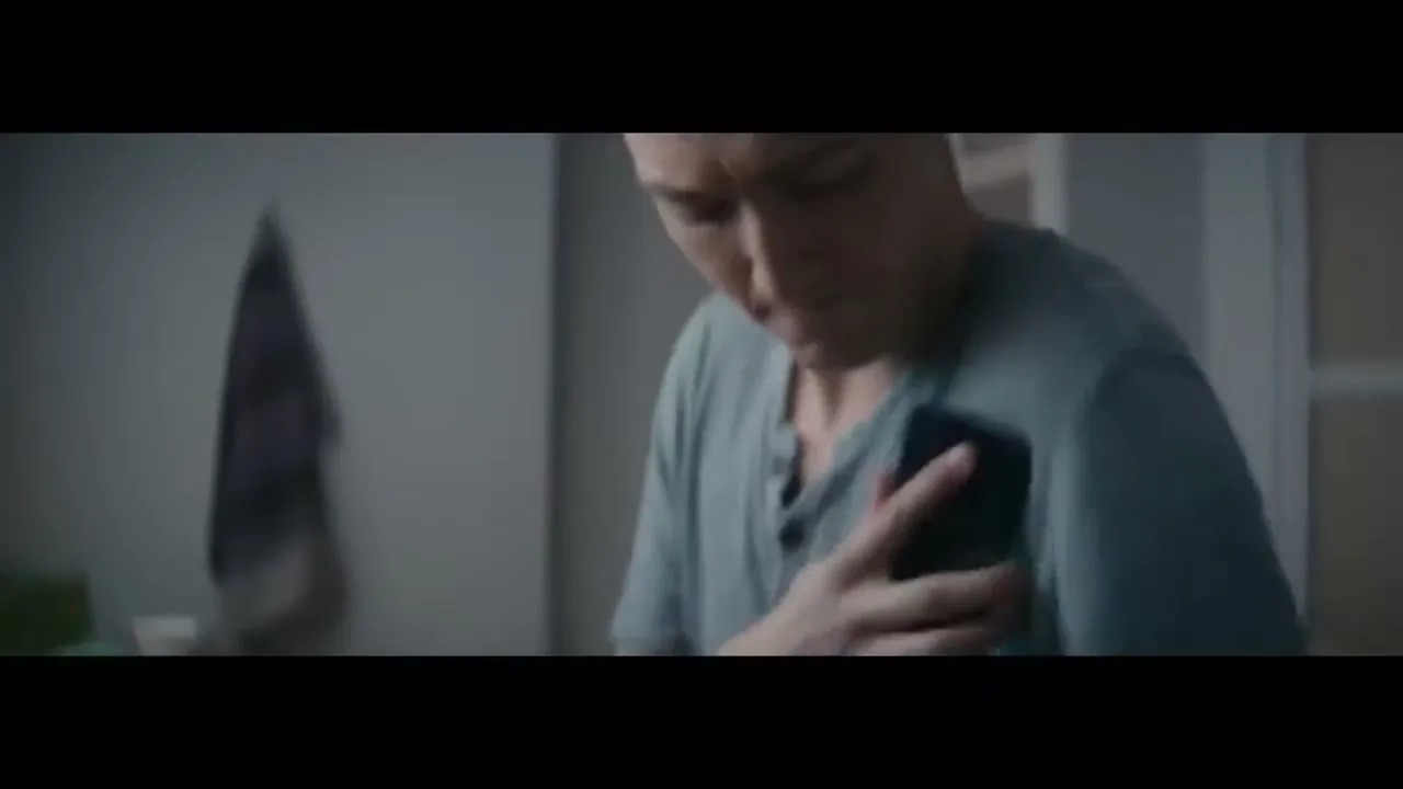 A young man in a blue shirt looks down at his phone, which he is wiping with his shirt, in a bathroom.