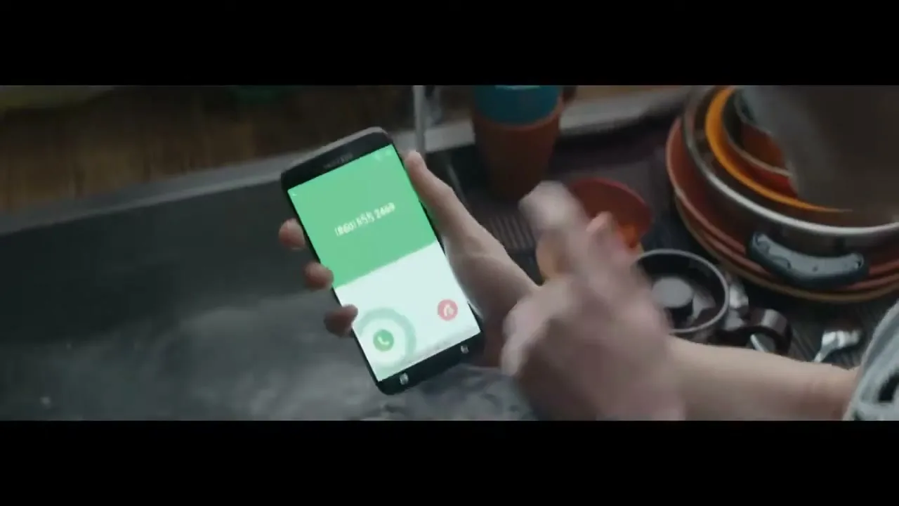 A person's hand holds a Samsung phone displaying an incoming call, then drops it into a sink full of water and dirty dishes.