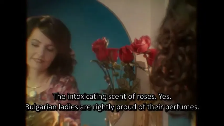 A woman in her 30s with dark hair and a floral dress looks at her reflection in a mirror, smiling, with red roses in the foreground.
