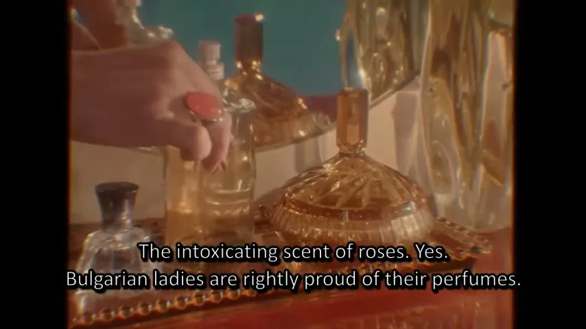 A woman's hand with a coral ring picks up a perfume bottle from a vanity tray with other ornate bottles and a mirror.
