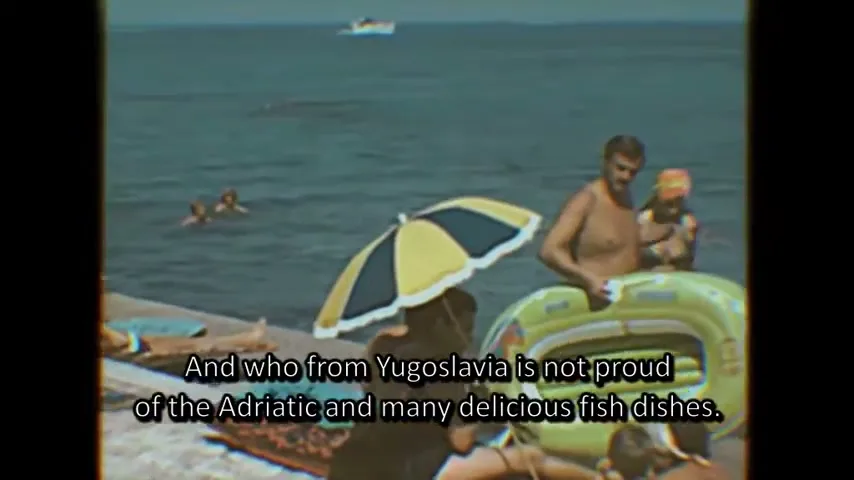 A man in swim trunks stands by the water with an inflatable ring, surrounded by other people on a sunny beach.
