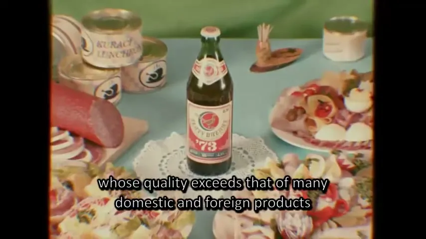 A bottle of Zlaty Bazant '73 beer stands on a white doily amidst various canned and prepared foods on a light blue tablecloth, with a vintage filter.