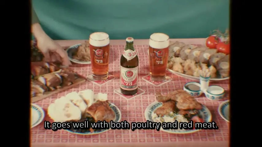 A table laden with food, including roasted chicken, bread, and other dishes, with a bottle of Zlaty Bazant 73 beer and two full glasses of beer in the center.