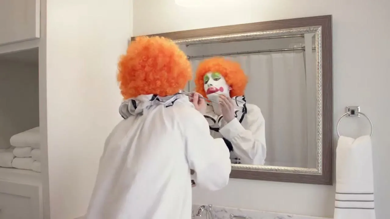 A person in a clown costume with an orange wig applies makeup to their face while looking into a bathroom mirror.