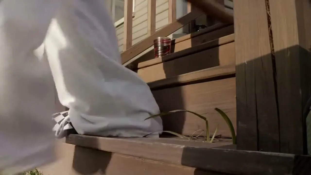 A person wearing white pants and black and white checkered shoes walks up wooden porch steps, with a red can visible on a higher step.