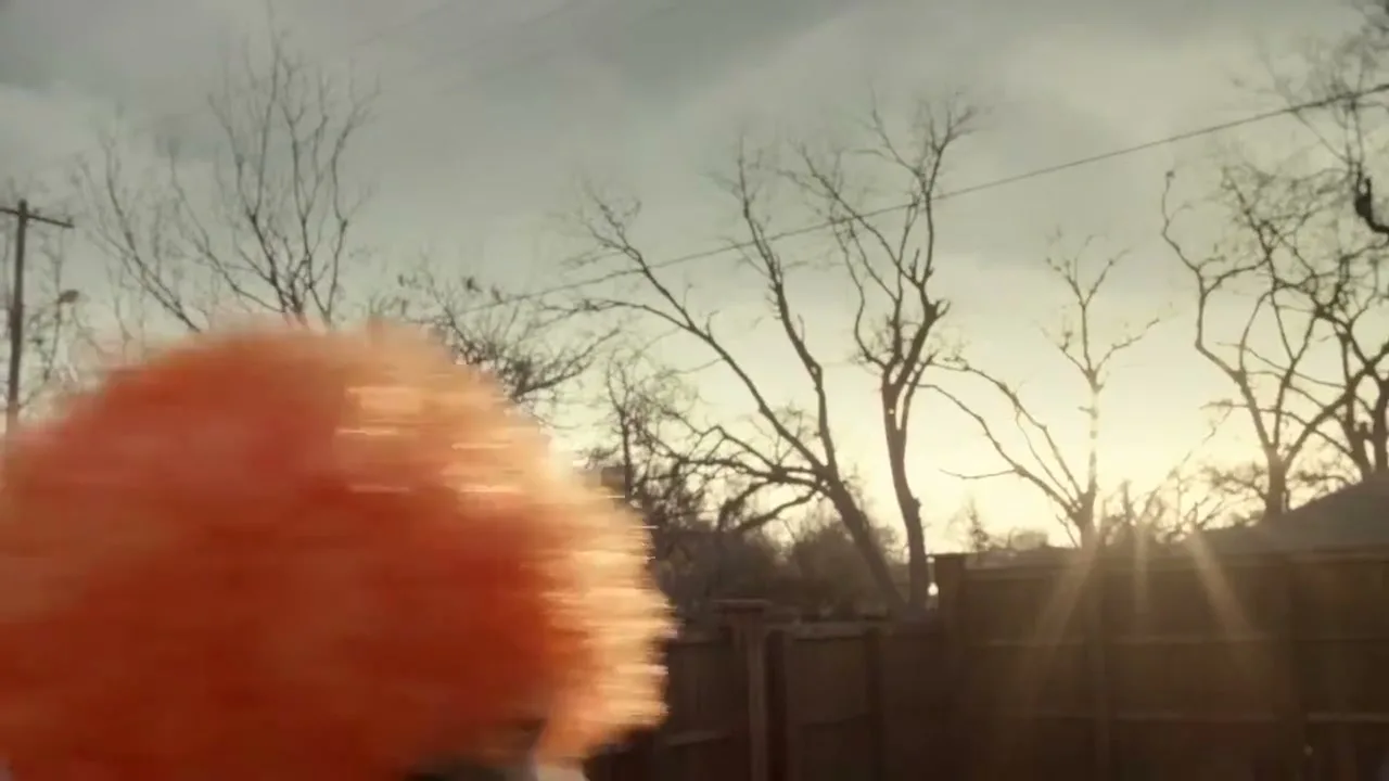 A bright orange, fluffy object quickly moves across the frame from left to right, with a wooden fence and bare trees in the background under a cloudy sky.