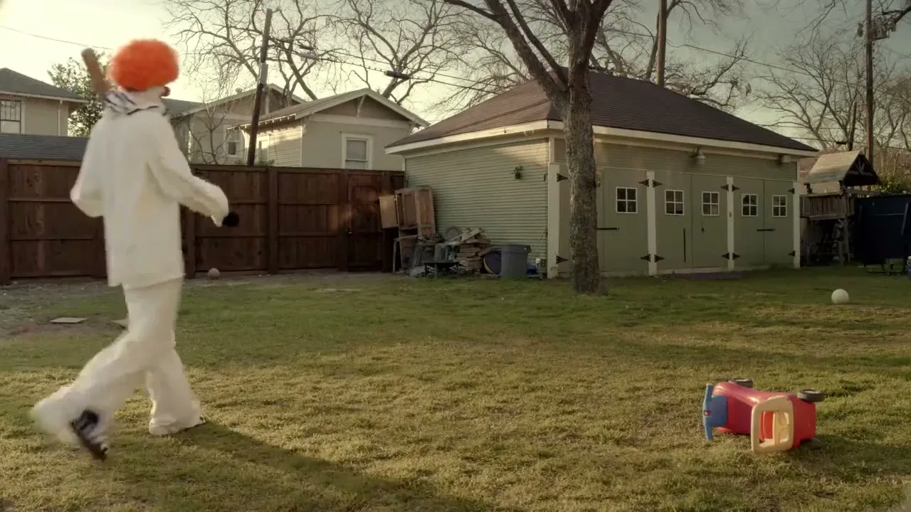 A person in a white clown costume with an orange wig walks across a grassy backyard towards a shed, holding a baseball bat.