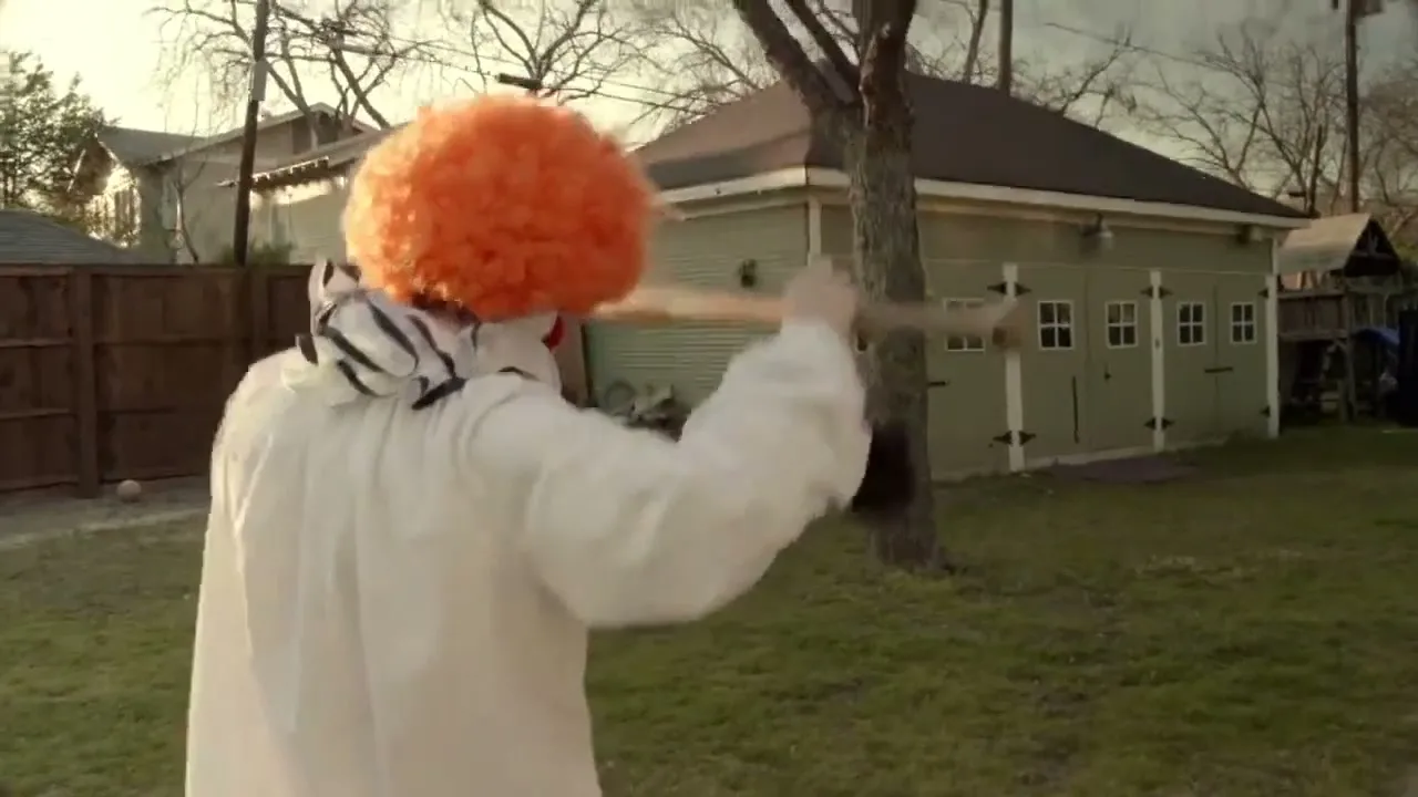 A person in a clown costume with an orange wig swings a baseball bat in a suburban backyard, facing away from the camera.