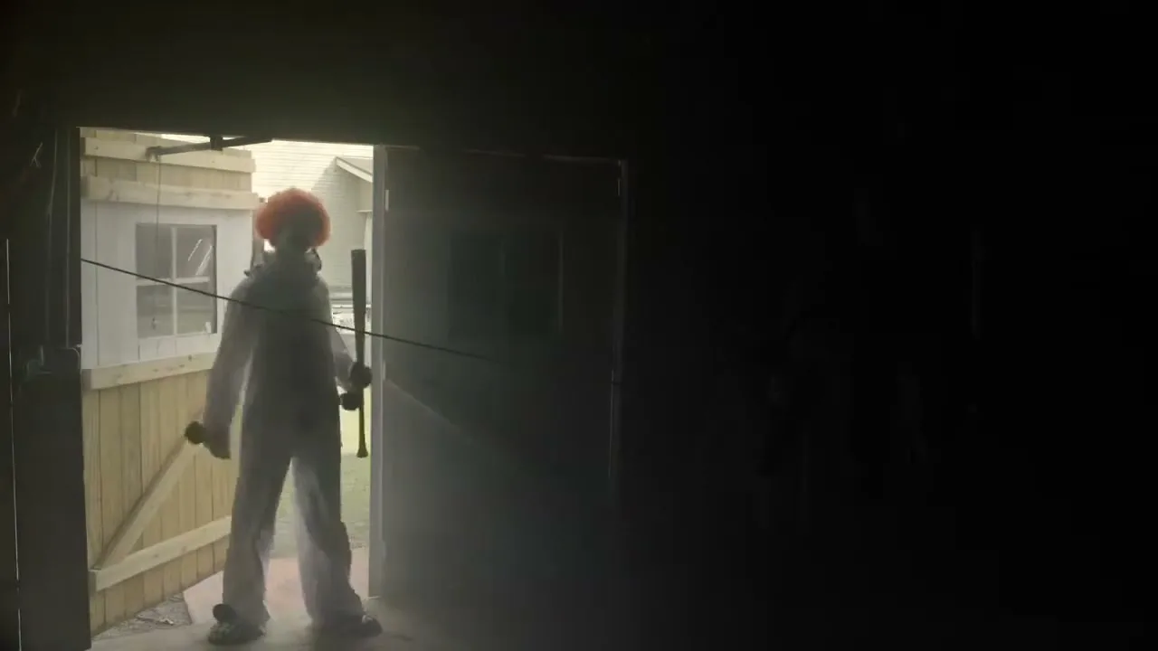 A person dressed as a clown with an orange wig and white jumpsuit stands in a doorway, holding a baseball bat, looking out into a bright exterior.