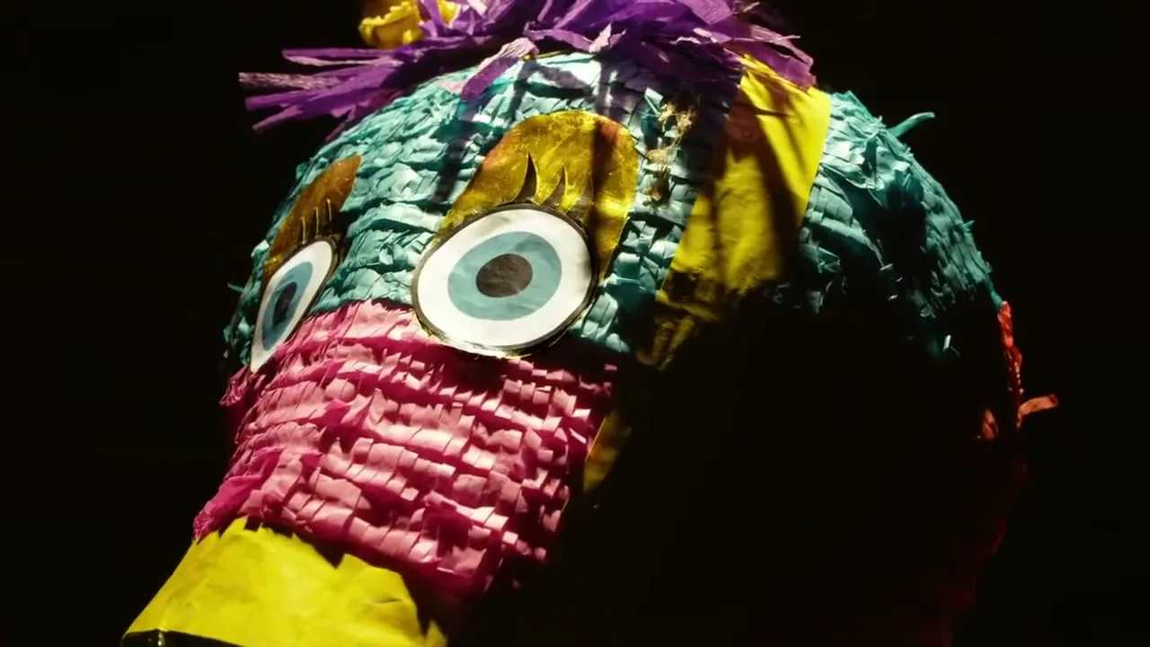 A colorful piñata, shaped like a bird with large eyes and a yellow beak, is shown in a dark setting.