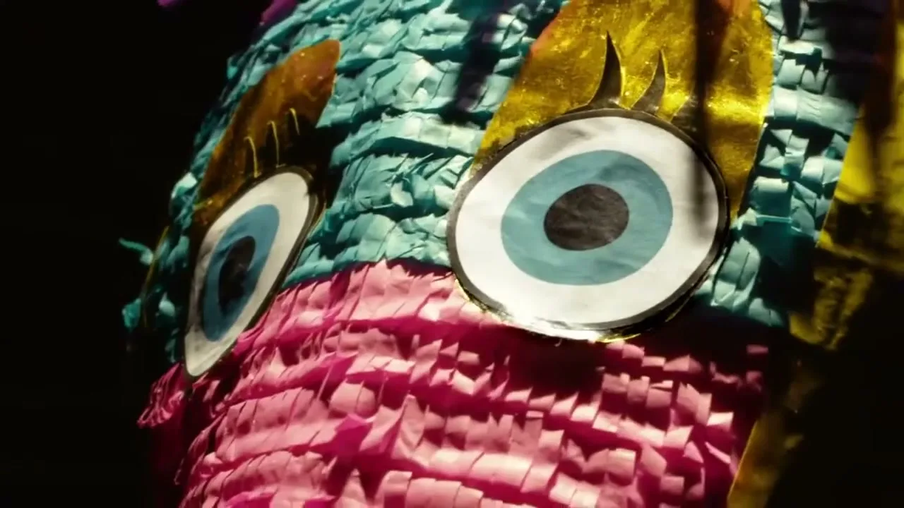 A colorful piñata with large, wide eyes is shown in a medium closeup, slightly moving against a dark background.
