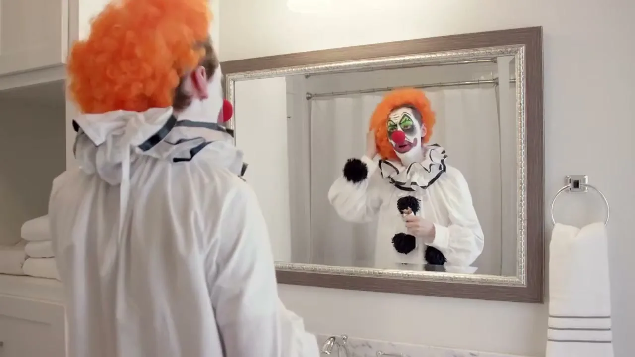 A person dressed as a clown with an orange wig and full makeup looks at their reflection in a bathroom mirror, adjusting their wig.