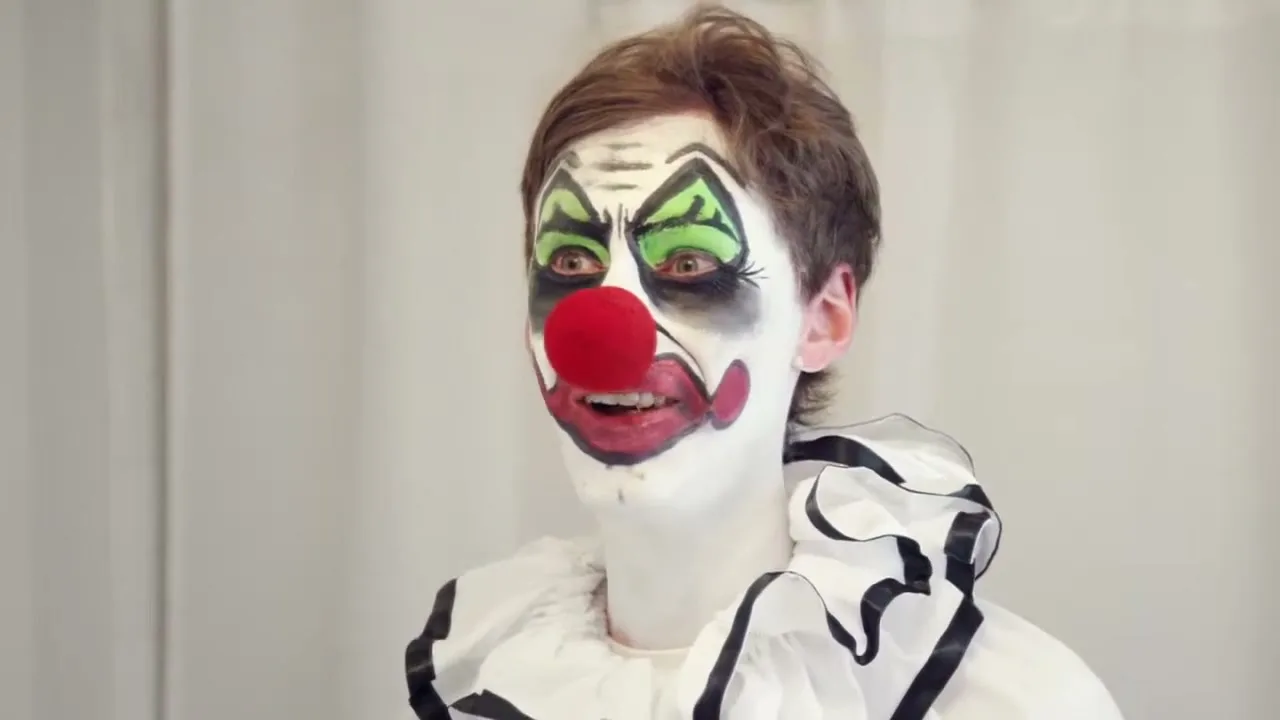 A young person with clown makeup and a ruffled collar laughs maniacally, looking directly at the camera in a brightly lit room.