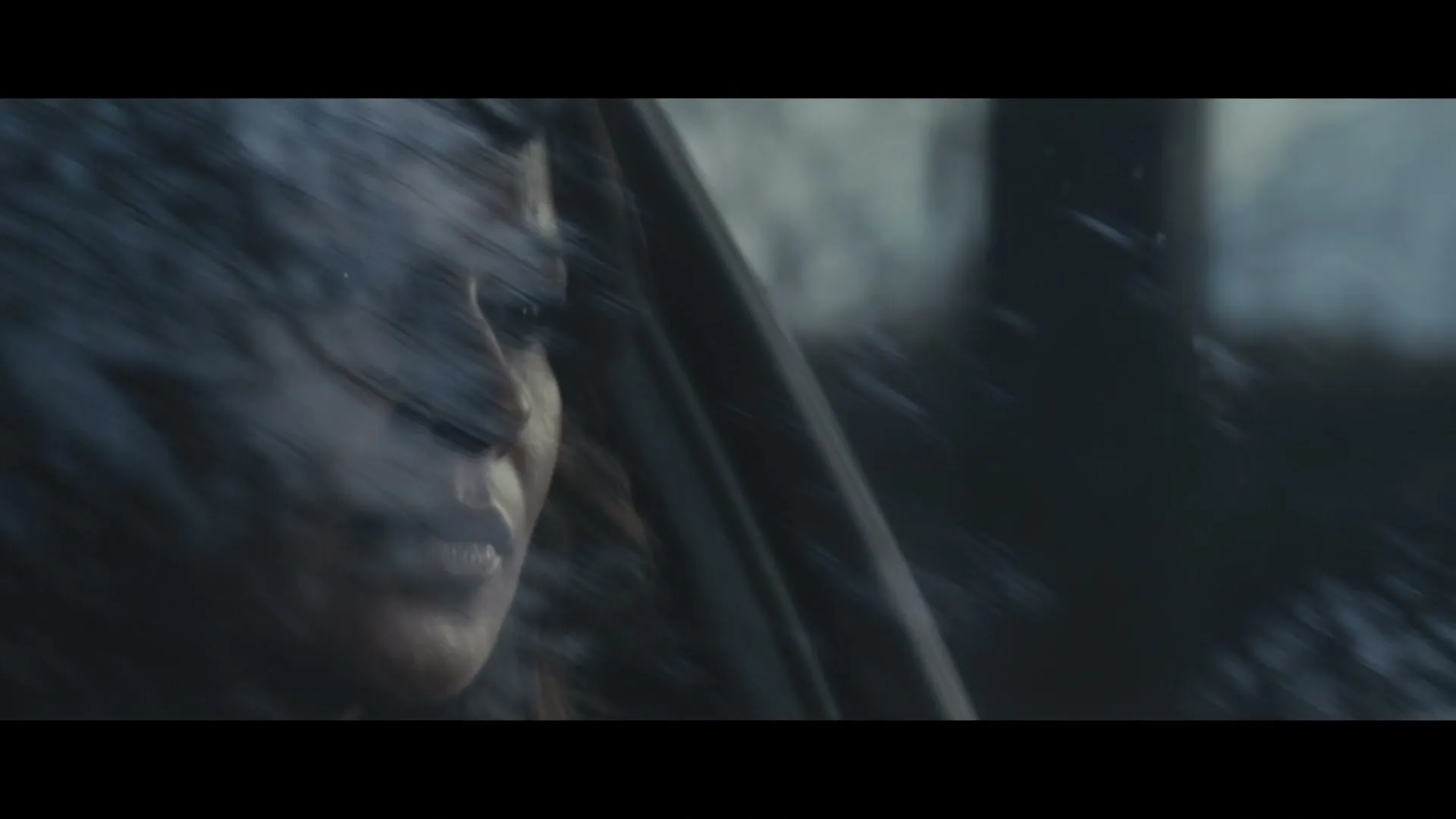 A woman with dark hair and a dark scarf looks out of a car window, with reflections of trees on the glass.