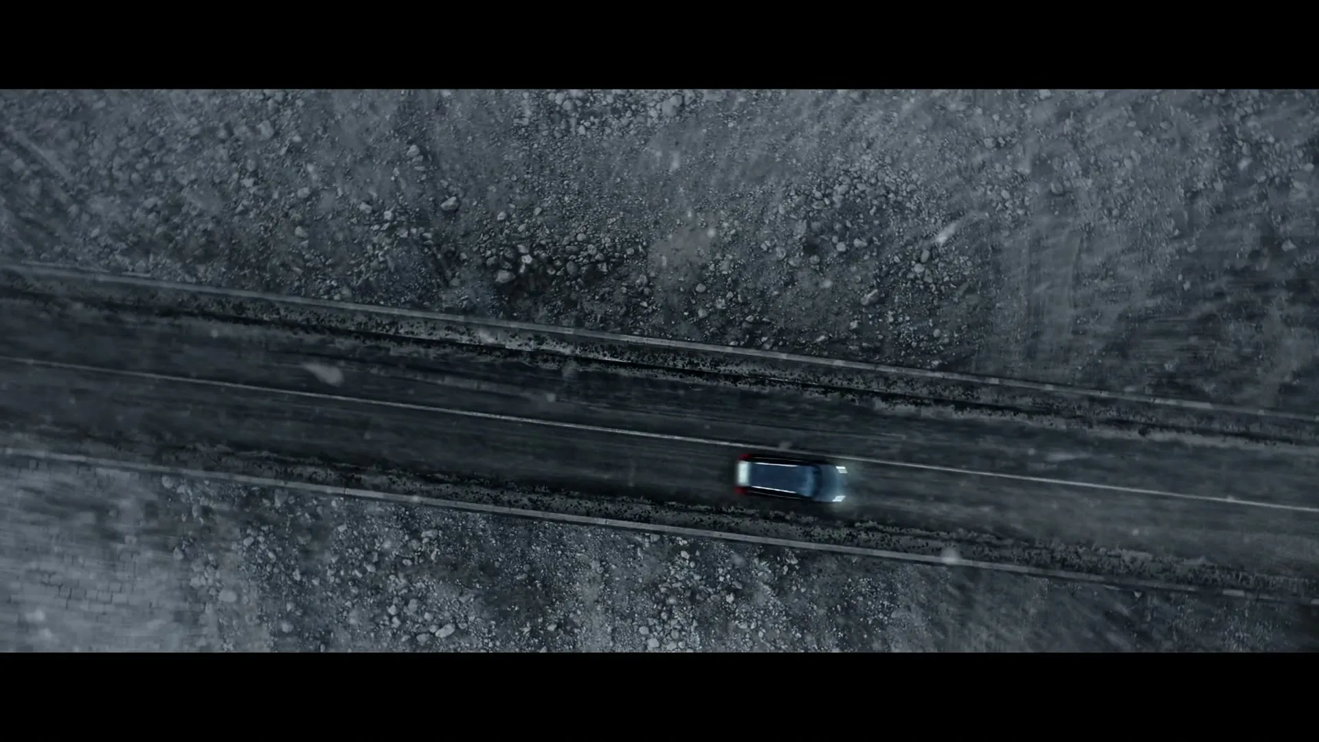 An aerial shot shows a dark car driving on a snowy, icy road through a desolate, rocky landscape during the day.