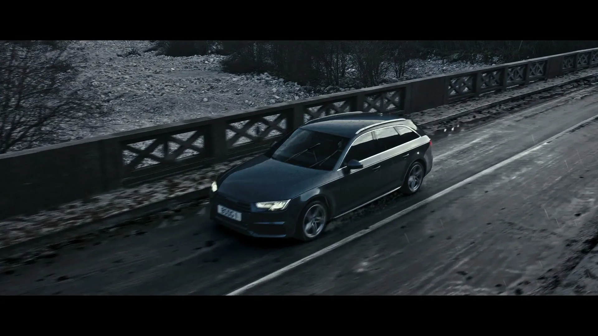 A dark grey station wagon drives on a wet, dark road over a bridge with a rocky riverbed below, under a dark sky.