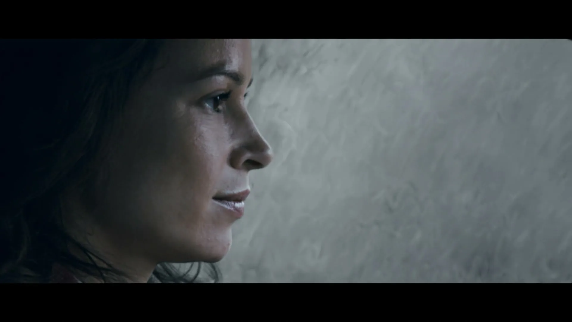 A woman with dark hair looks out a car window at the rain, her face illuminated by soft light.