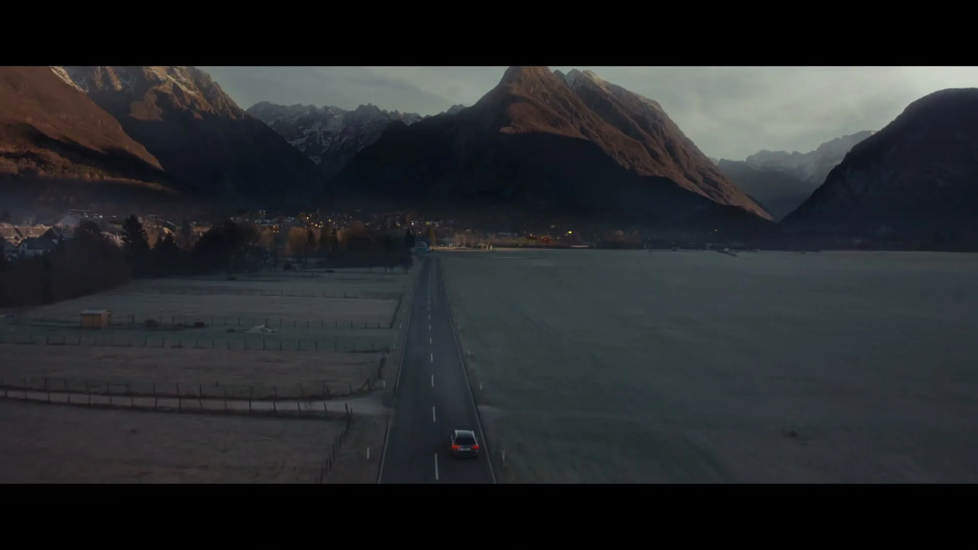 An aerial view shows a car driving down a long, straight road through a valley with snow-capped mountains in the background during golden hour.