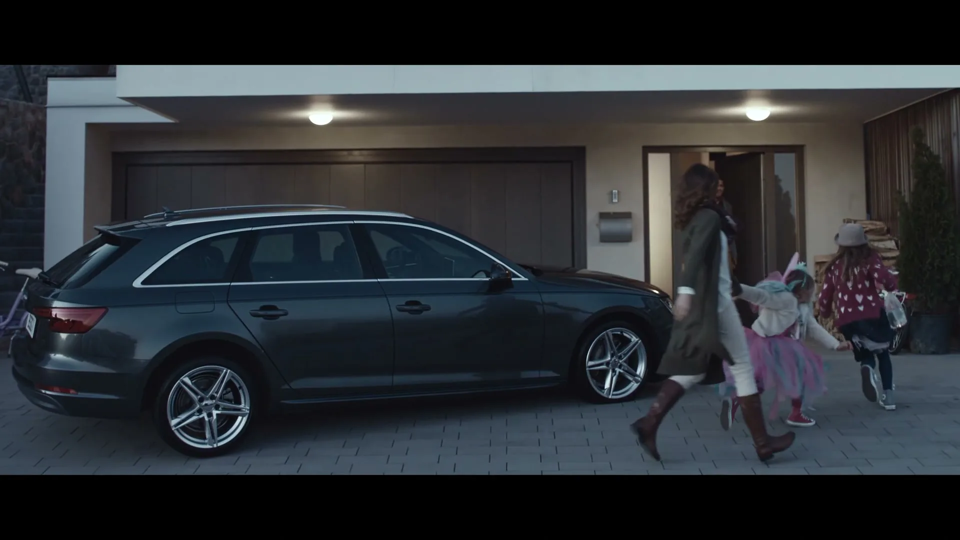 A family of four, a mother, father, and two young daughters, run out of their modern home towards a dark grey station wagon parked in the driveway.