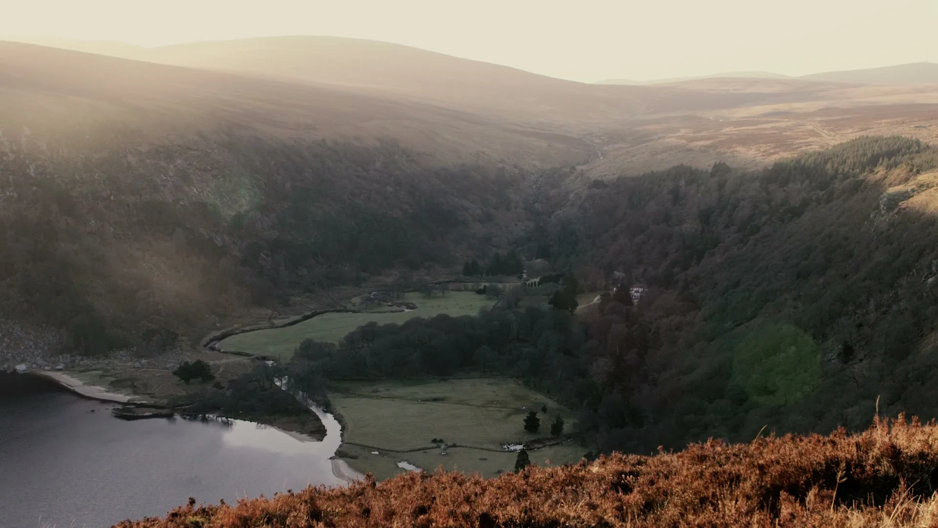 An extreme wide shot shows a vast, sunlit valley with a dark lake, a winding river, and green fields surrounded by hills and trees.