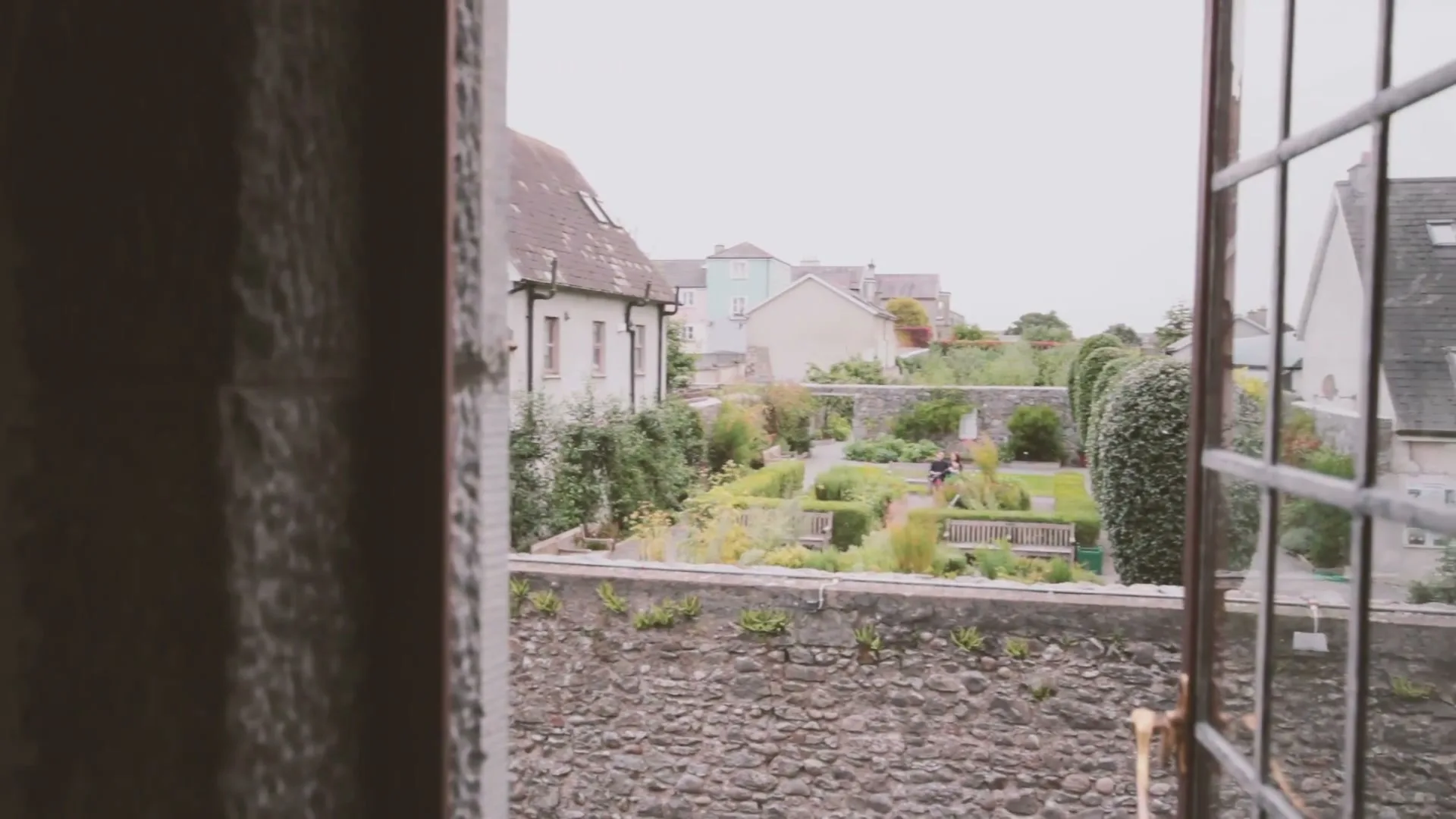 A wide shot from inside a window shows a person walking through a manicured garden with stone walls and old buildings under a bright sky.