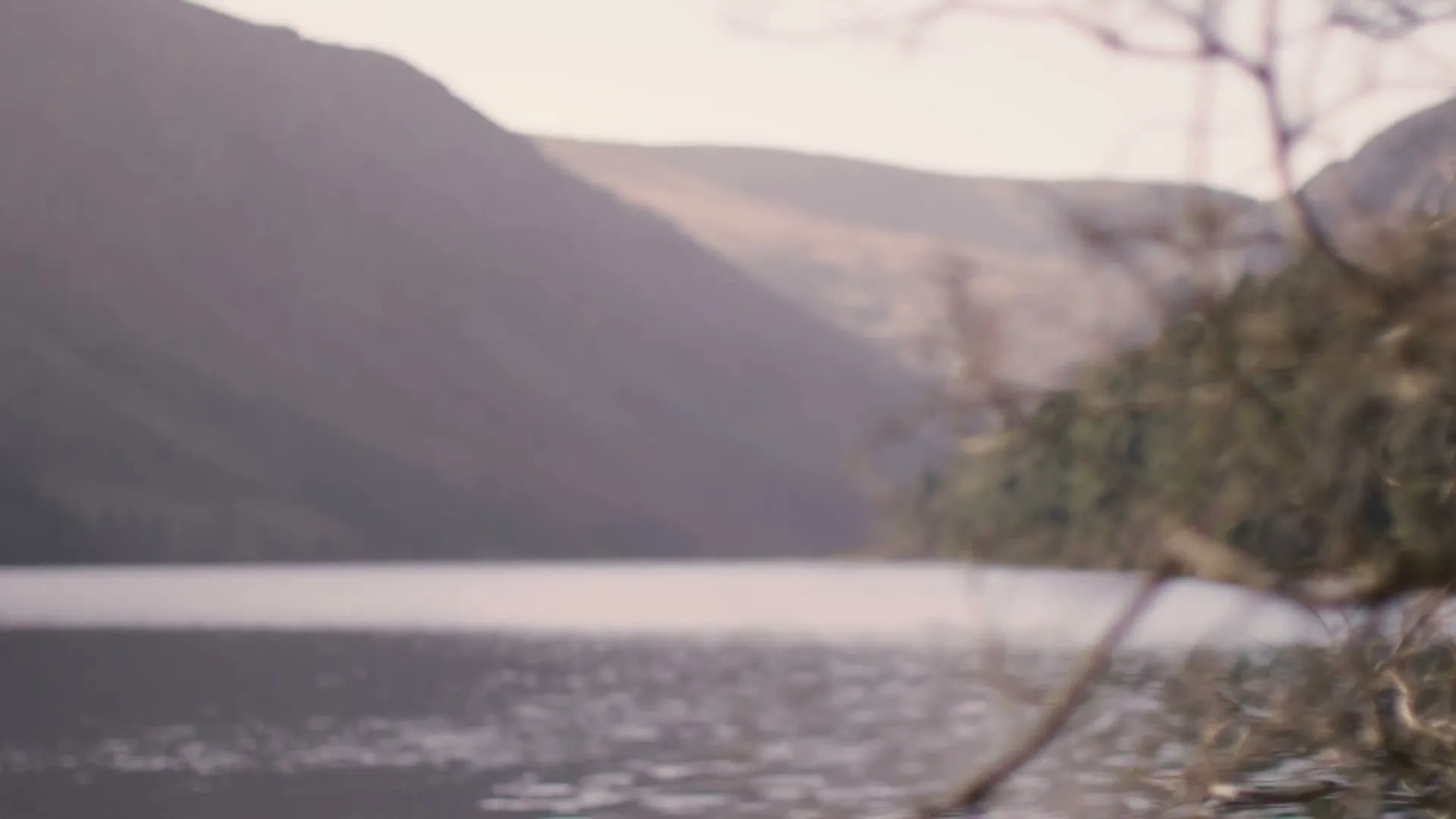 A wide shot of a lake surrounded by mountains, with bare tree branches in the foreground, as the camera slowly pans right.