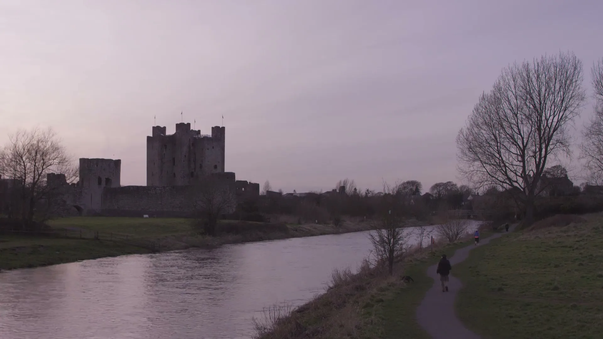 A wide shot of a medieval castle by a river at dusk, with a person walking on a path in the foreground.