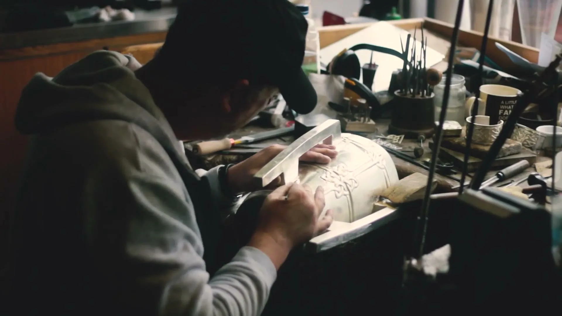 A male artisan in a grey hoodie and black cap meticulously engraves intricate patterns onto a metal vessel at a workbench in a dimly lit workshop.