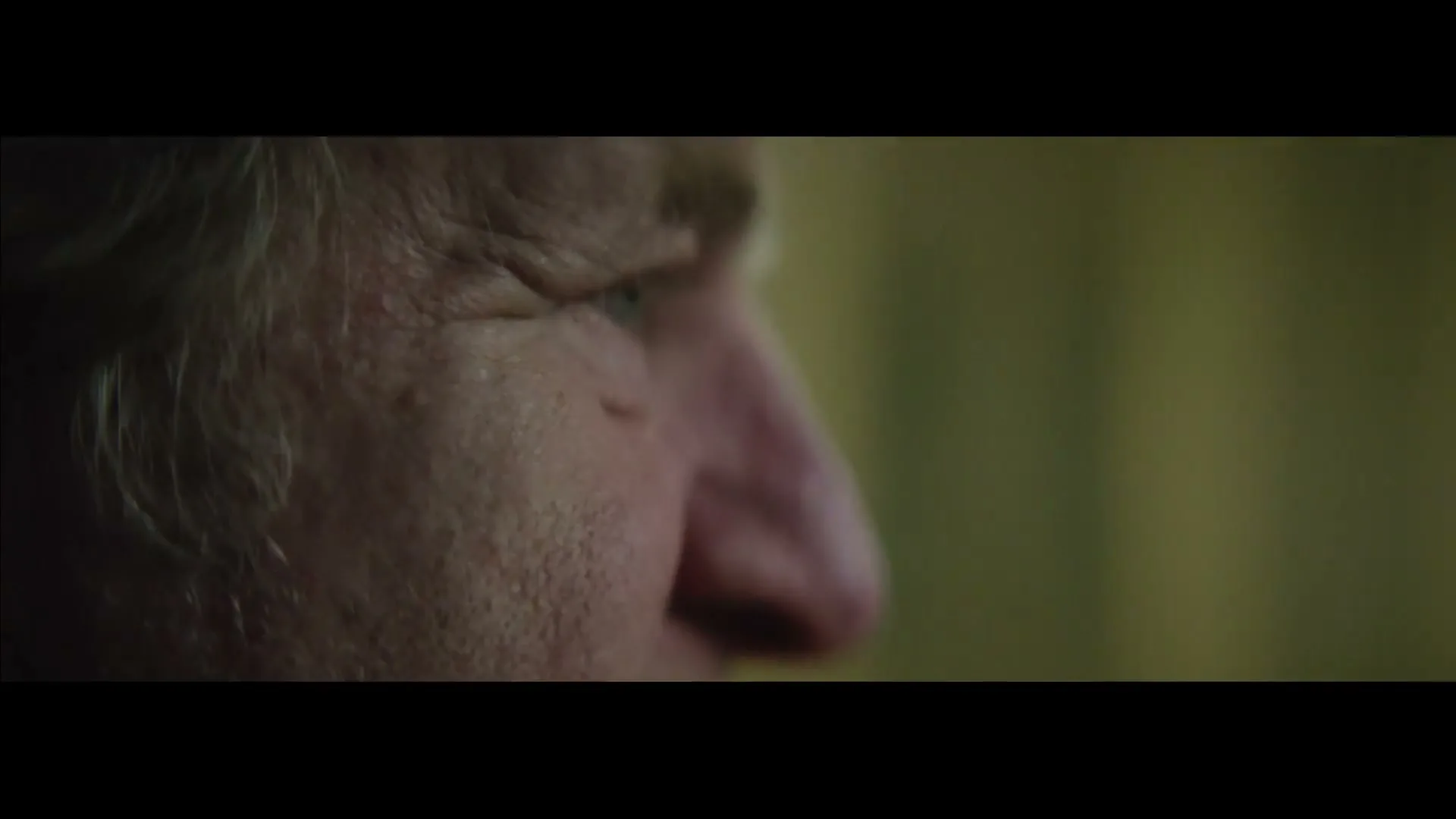 A man's face, with white hair and a prominent nose, is shown in profile, looking intently at something off-screen in a dimly lit room.
