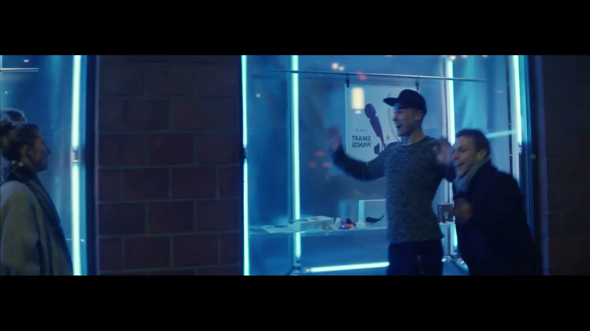 Two young men in casual clothes dance excitedly in front of a brightly lit store window at night, while two women watch them.