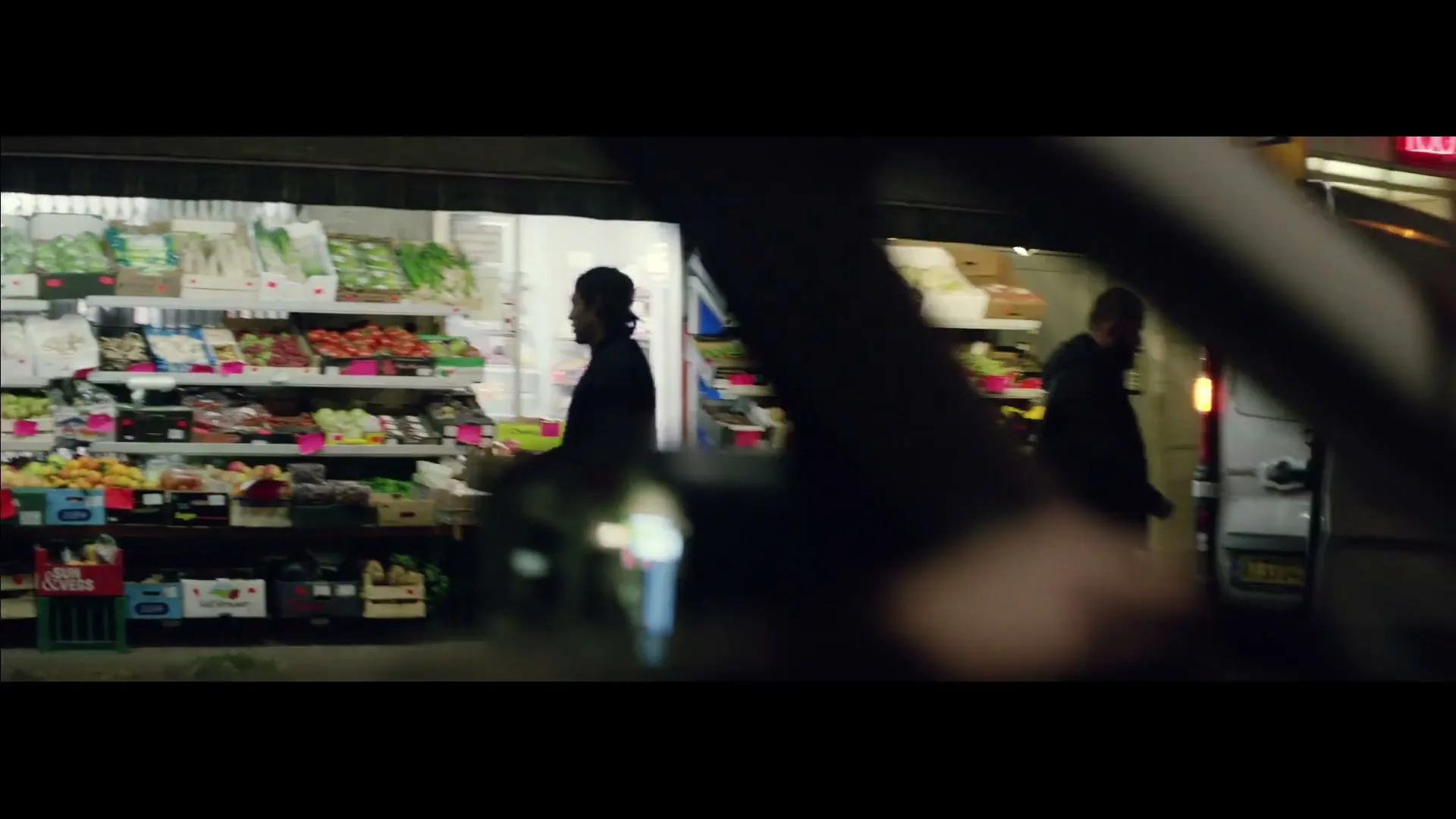 A car drives past a brightly lit grocery store and a food truck at night, with people walking on the sidewalk.