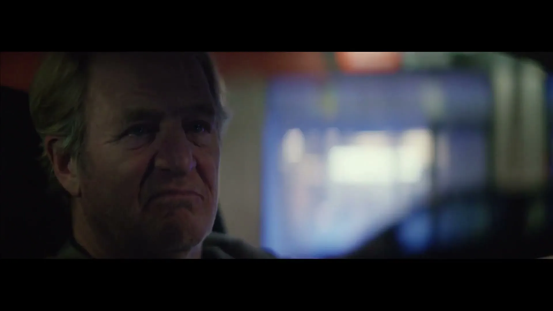 A man with light brown hair and a beard sits in a car at night, looking forward with a sad expression.
