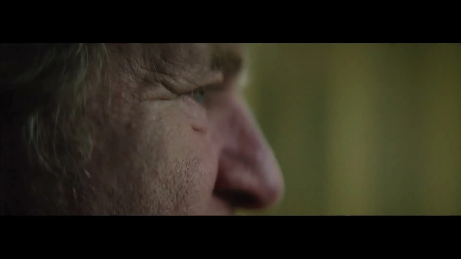 A blurry extreme closeup of an older man's face, looking off-screen, as the camera slowly racks focus to reveal his eye and nose in a dimly lit interior.