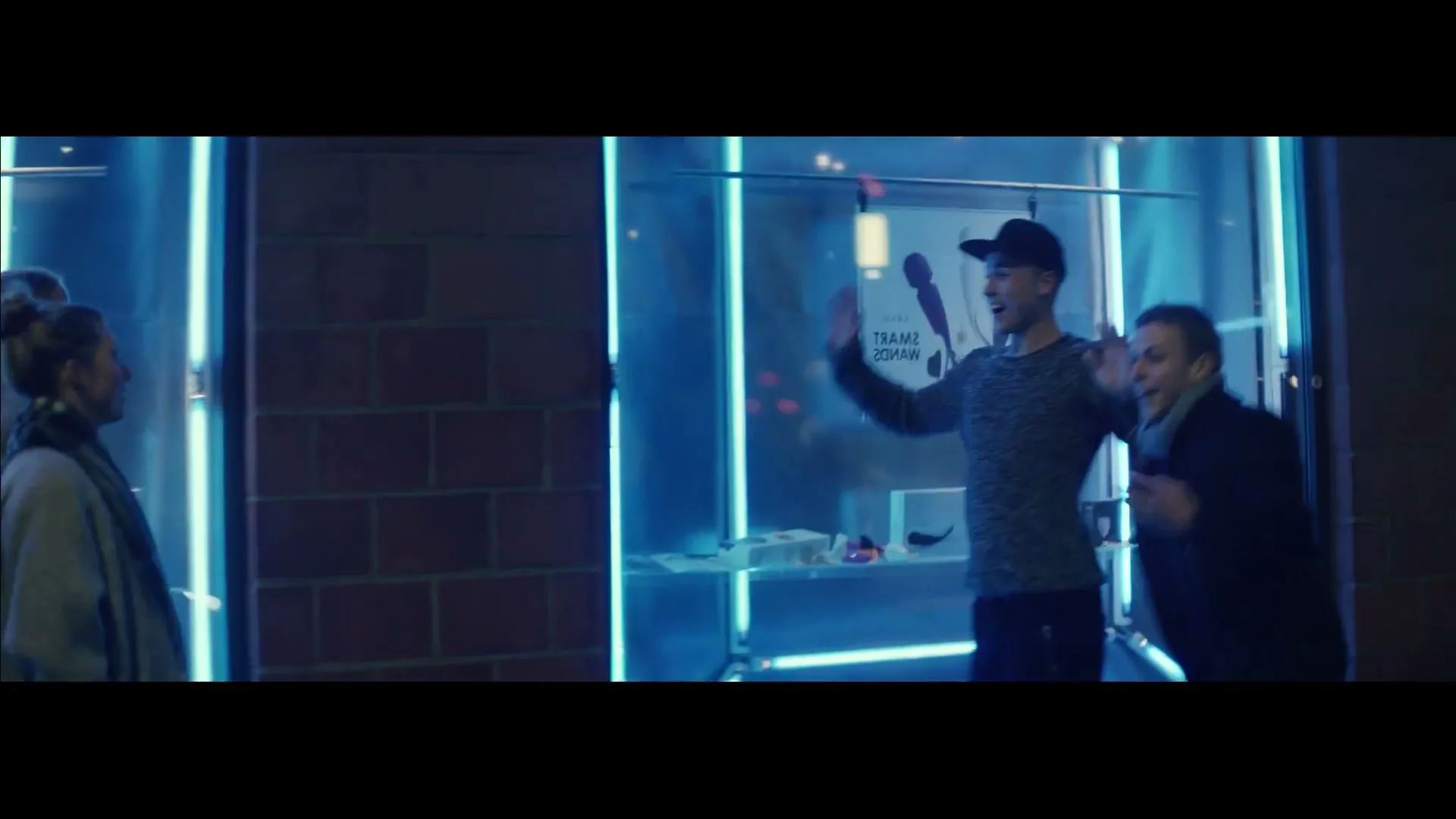 Two young men in casual clothes stand in front of a brightly lit store window at night, waving and smiling at two women walking by.