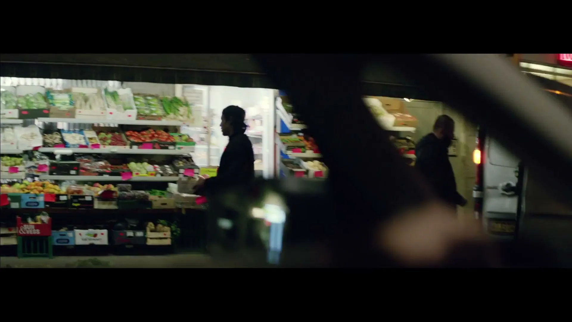 A car drives past a brightly lit grocery store at night, where several men are seen working and interacting outside the shop.