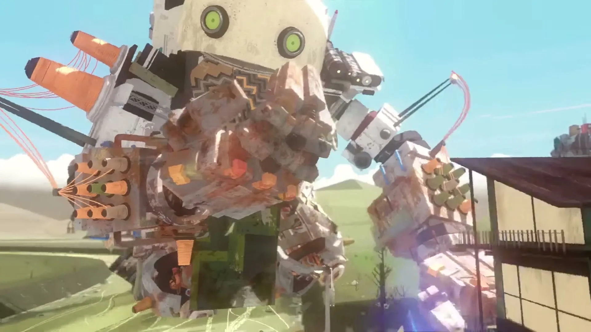 A giant, patchwork robot with green eyes and a zig-zag scarf stands in a field, as a group of people watch it from a wooden bridge.