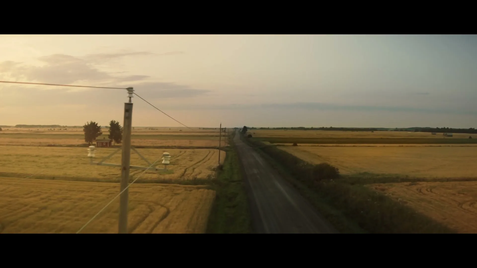 An aerial wide shot captures a long, straight road cutting through golden fields under a soft, cloudy sky at dusk, with a small red house and trees in the distance.