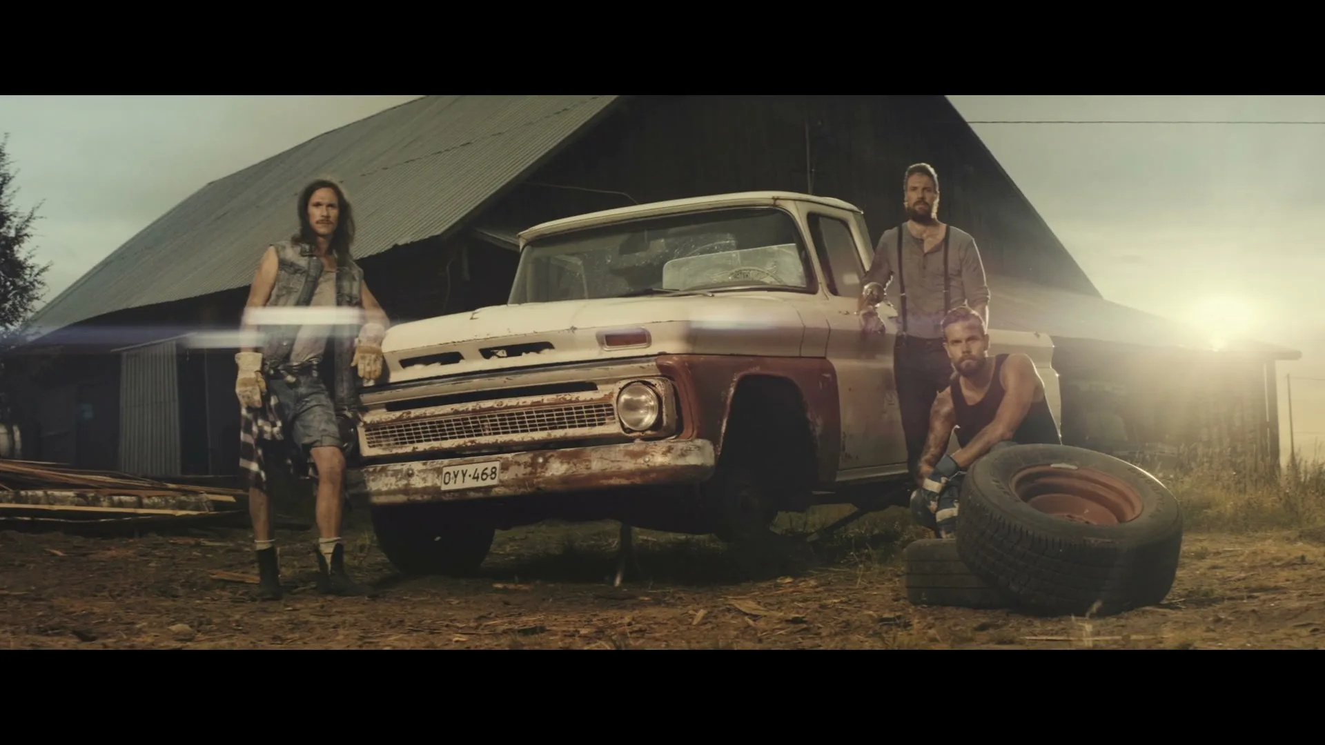 Three rugged men, dressed in casual workwear, pose next to an old pickup truck with a barn in the background, bathed in golden hour light.