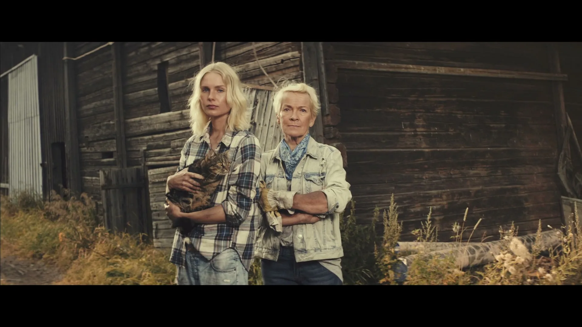 Two women, one holding a cat, stand side-by-side in front of a rustic wooden barn, looking directly at the camera with serious expressions.