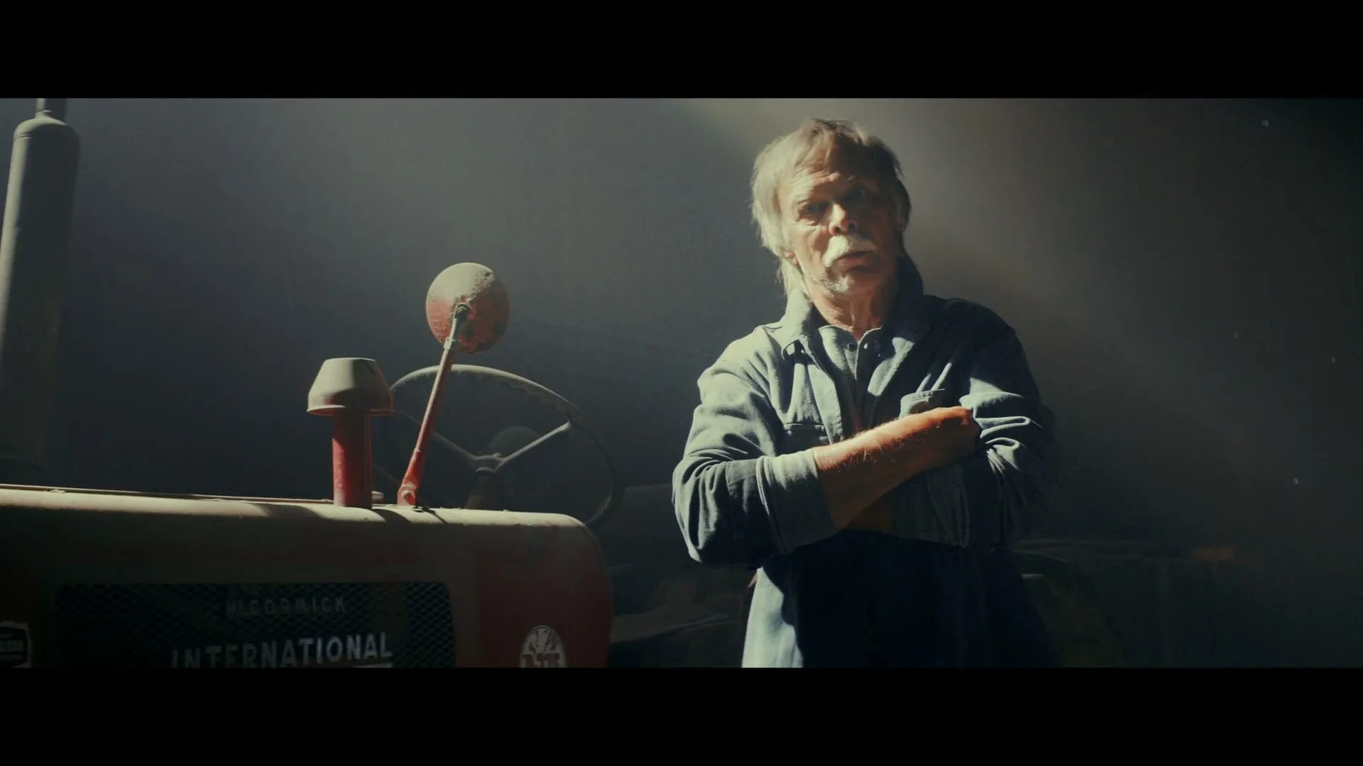 A weathered older man with a mustache stands with crossed arms next to a vintage red tractor in a dimly lit barn, looking directly at the camera.