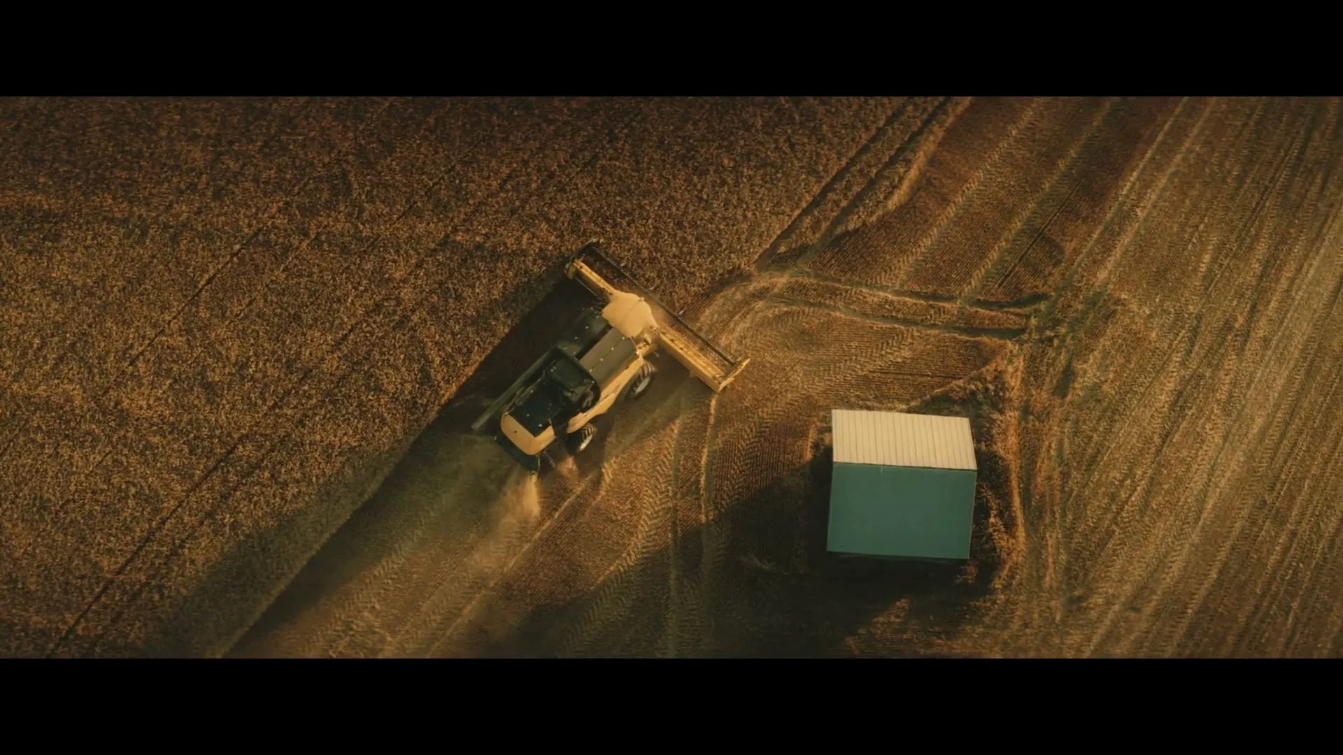 An aerial shot shows a combine harvester working in a field next to a small barn, creating dust trails under the golden hour sun.