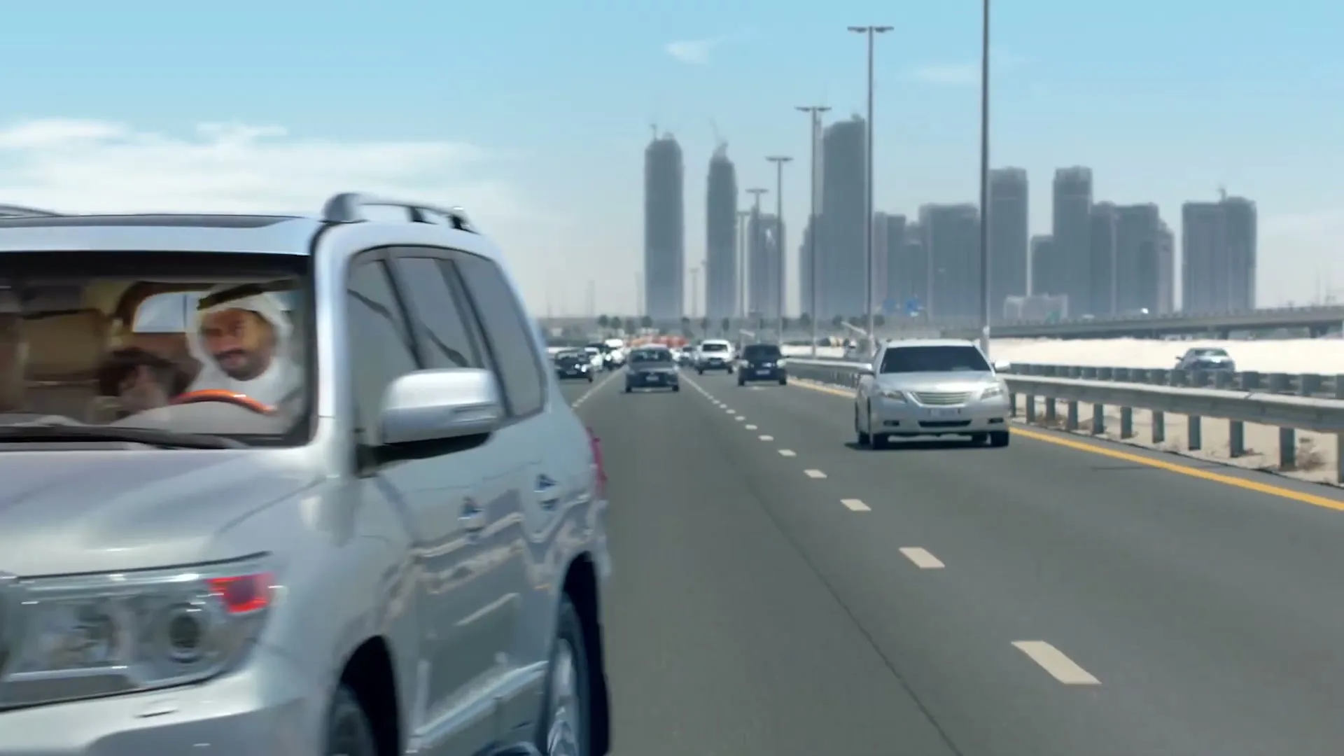 A family of four, dressed in traditional attire, drives in a silver SUV on a highway with a city skyline in the background, looking happy.