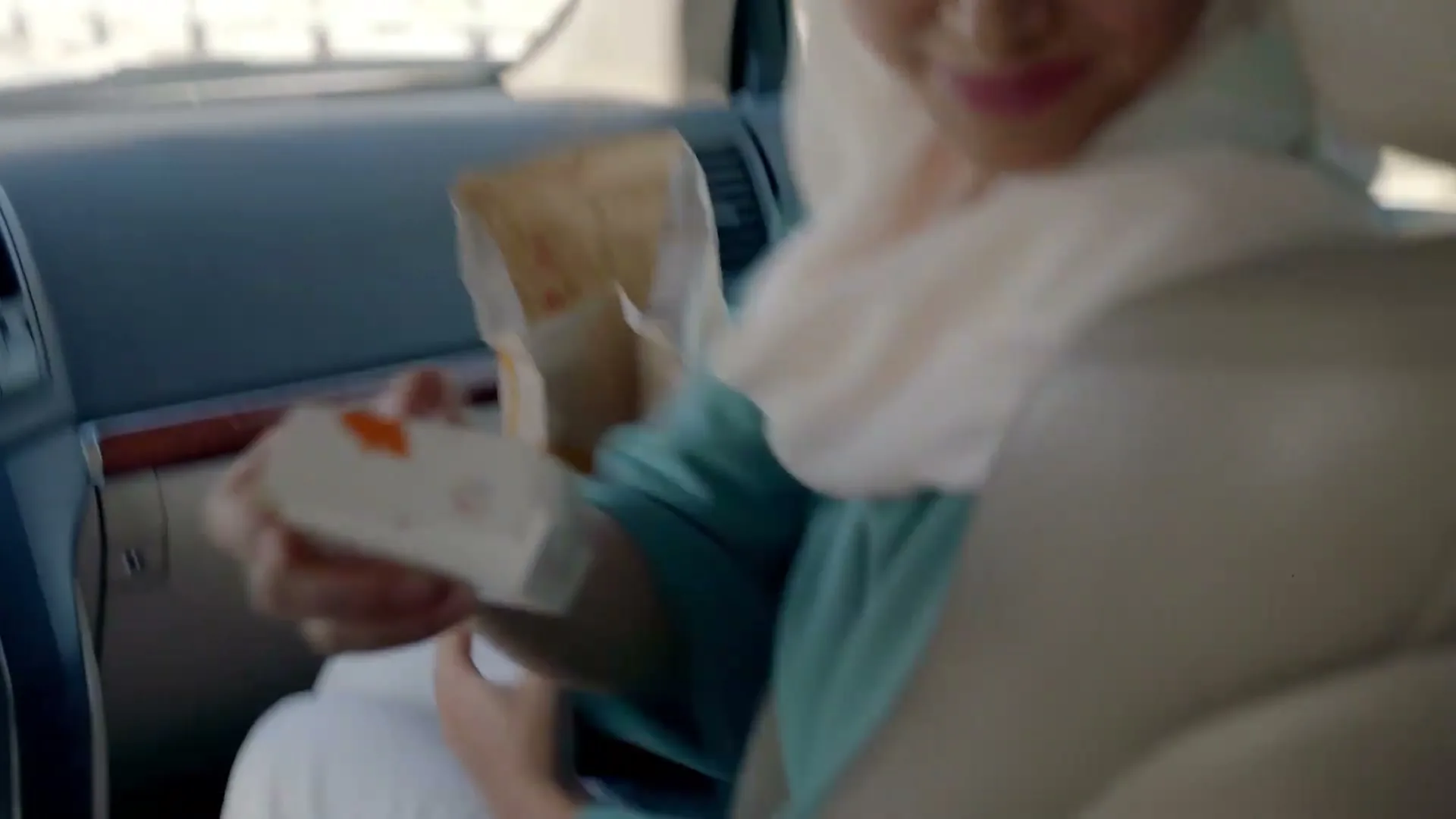 A woman in a hijab passes a McDonald's Happy Meal box to a child in the back seat of a car, driving on a highway.