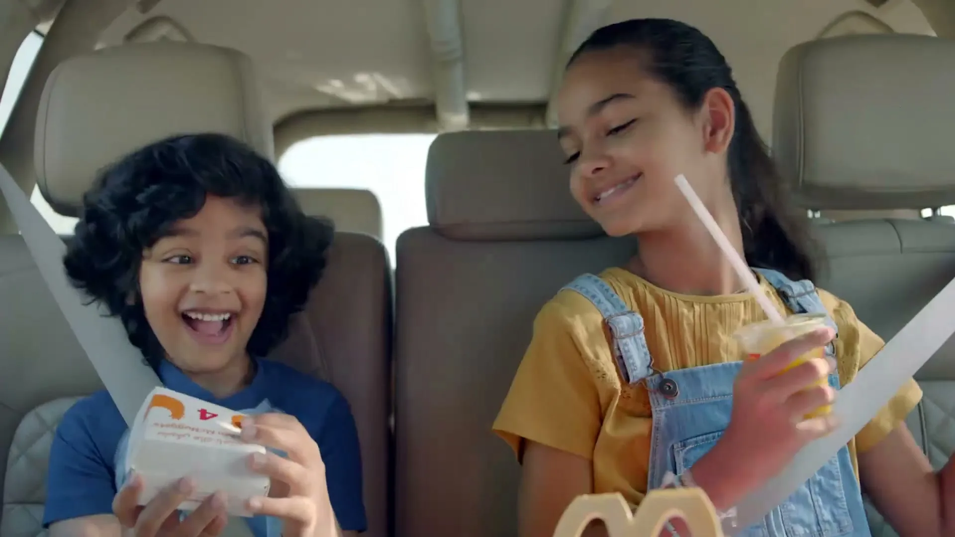 Two happy children, a boy and a girl, are sitting in the back seat of a car, holding McDonald's food and drinks.