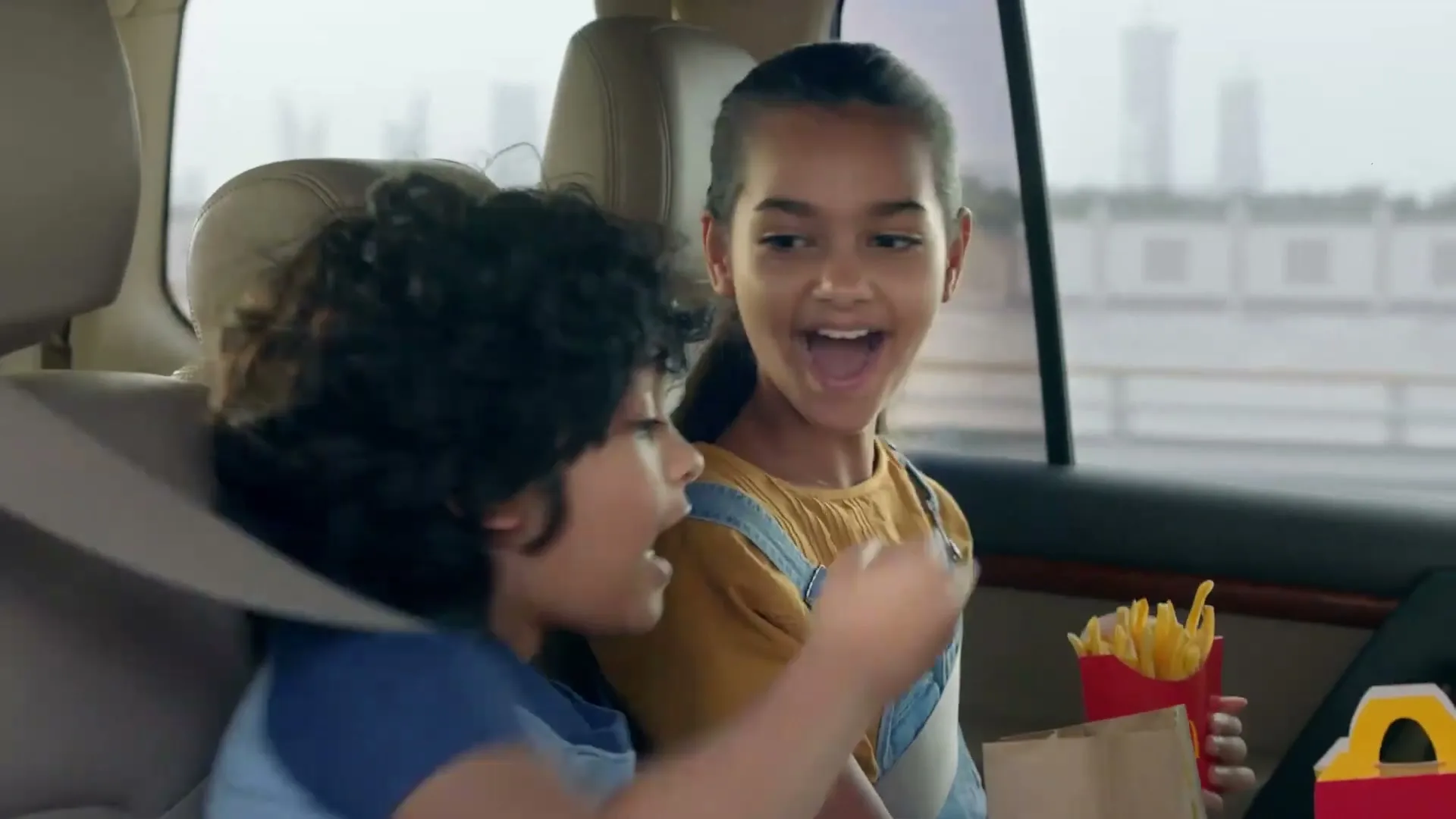 A young girl in a car offers a french fry to a young boy, both smiling and laughing in the back seat of a car.