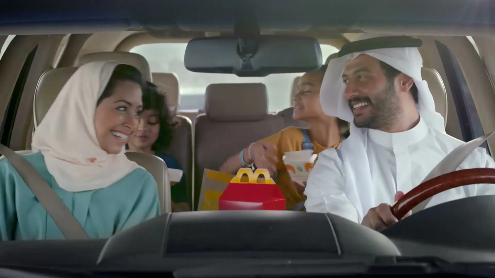 A family of four, dressed in traditional Arab attire, smiles and laughs in their car, with McDonald's Happy Meals on the back seat.