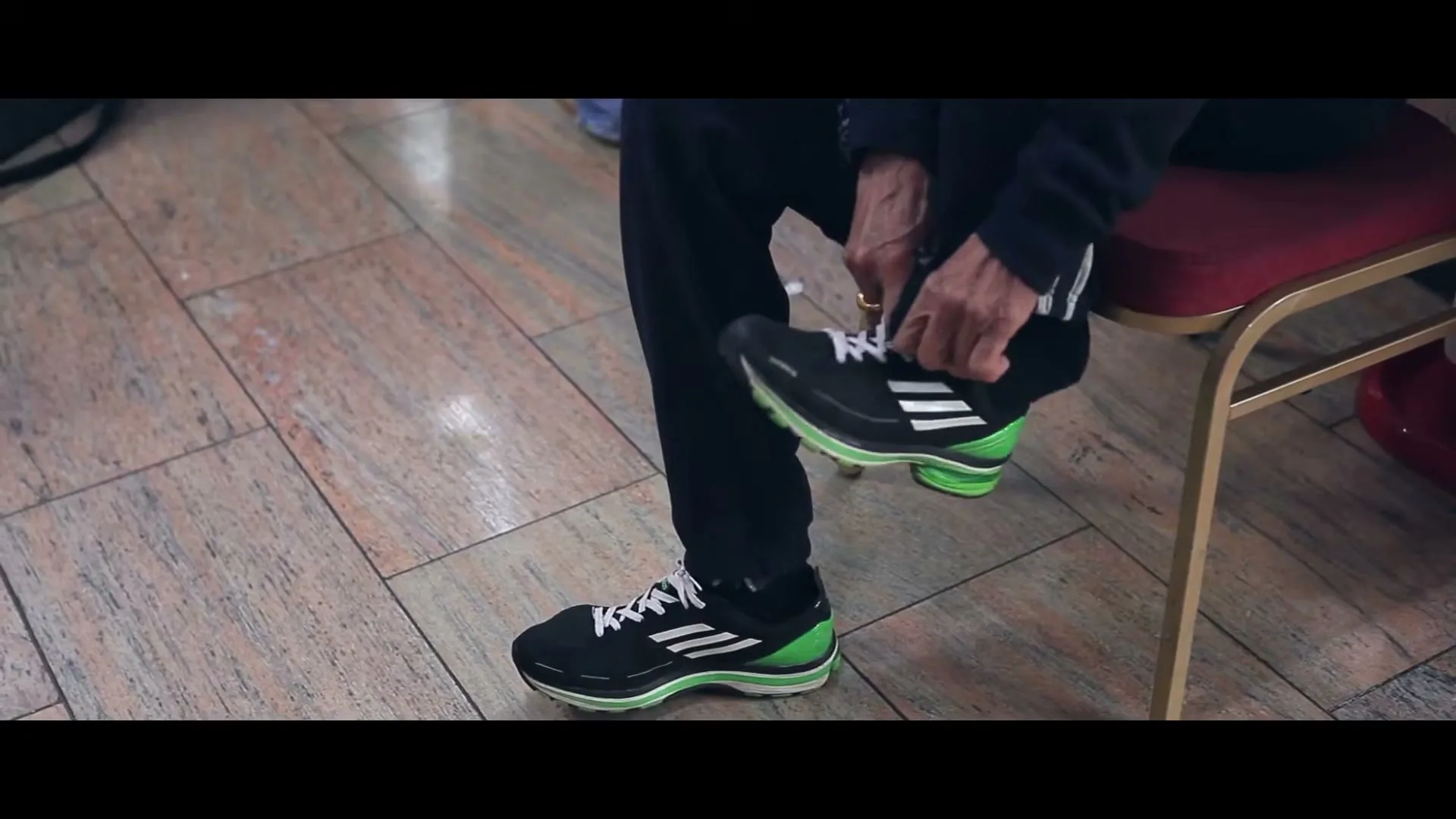 An elderly person with dark pants and black and green running shoes sits on a red chair, tying their shoelaces on a tiled floor.
