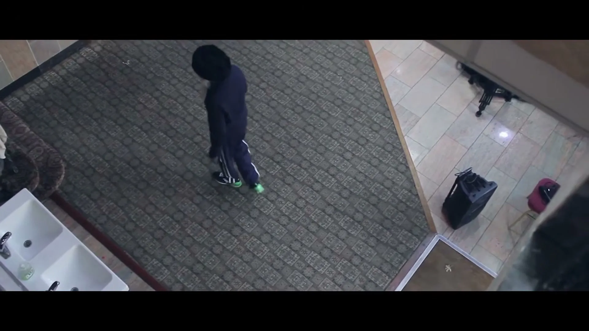 A person in a dark tracksuit and black head covering walks across a patterned rug in a room with tiled floors, seen from an overhead perspective.