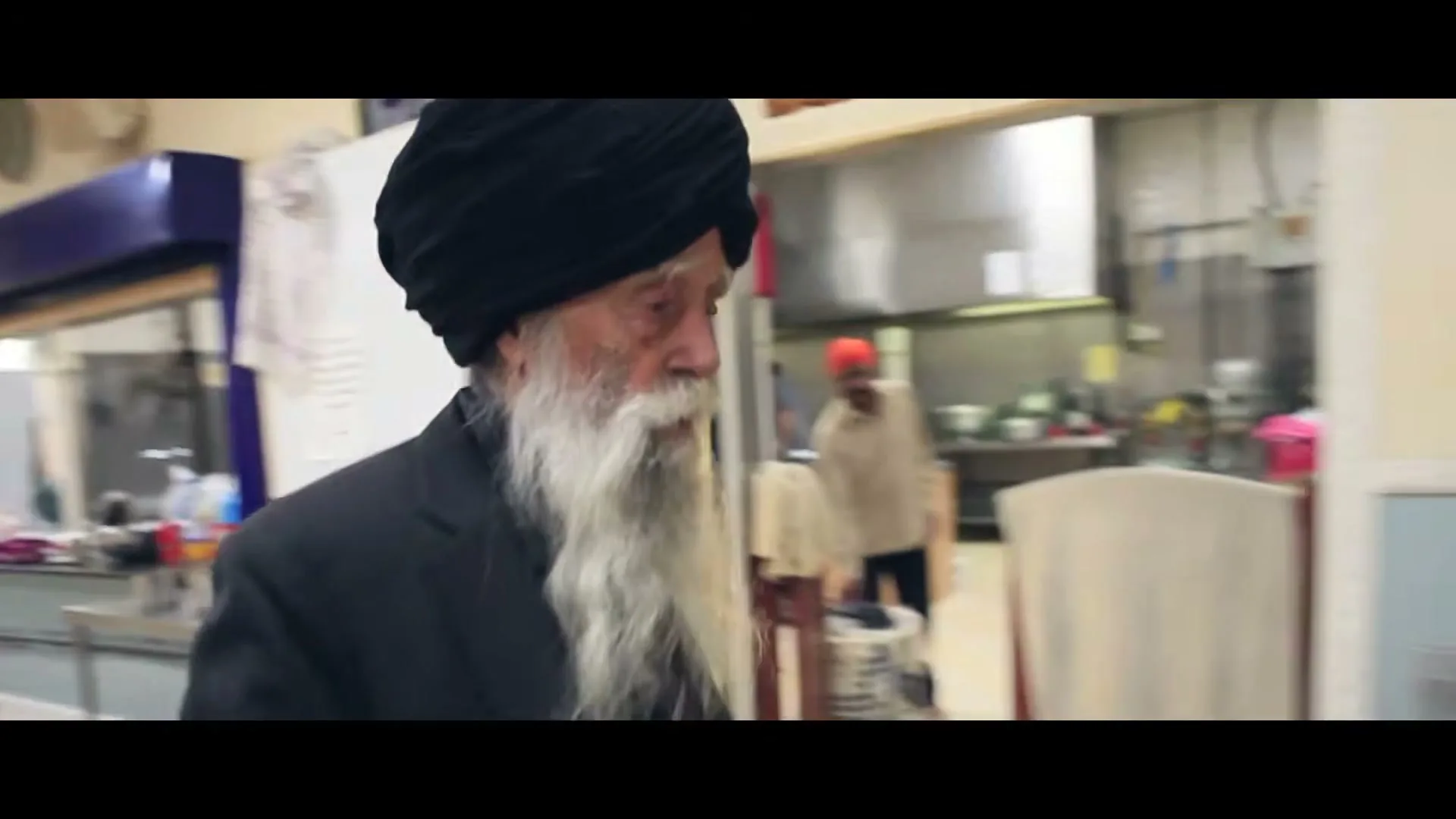 An elderly Sikh man with a long white beard and black turban walks through a community kitchen, then stops to prepare a drink.