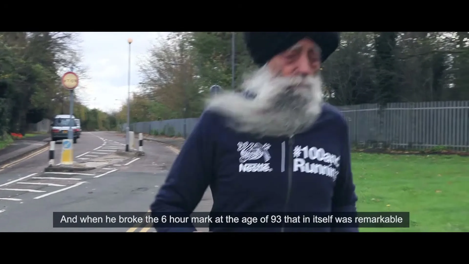 An elderly Sikh man with a long white beard and black turban runs on a paved road next to a grassy area on an overcast day.